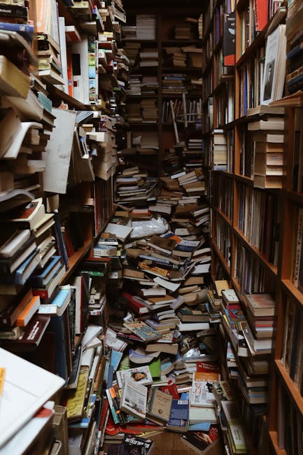 Inside a small, cluttered space with wooden bookshelves on both sides, densely packed with books, magazines, and papers. Some books are stacked horizontally or leaning, creating a disorganized appearance. The floor is covered with a mixture of loose books, open publications, and packaging materials, including cardboard and paper packaging. A doorway leading outside or into another room is partially visible at the far end, with some boxes and items placed nearby, indicating recent movement or packing activity. The environment appears to be part of a home undergoing a relocation process or clearing, with items ready for collection or disposal. The scene is lit by ambient light, highlighting the chaotic arrangement of packing and clutter, consistent with the process of home removals or furniture transport, as handled by [COMPANY_NAME].