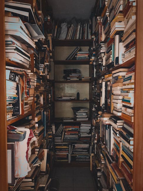 A narrow storage closet filled with numerous shelves stacked with a large collection of books, magazines, and documents. The books are arranged in various orientations, some standing upright, others stacked horizontally, with different sizes and colors. There are cardboard boxes and plastic storage containers positioned at the bottom and sides of the shelves, some partially open revealing packing materials like paper and bubble wrap. The shelves are made of metal and wood, and the space is dimly lit, likely illuminated by a small overhead light. The floor is covered with dark tiles, and a partial view of an adjacent doorframe is visible on the right side. The dense arrangement of items suggests it may be used for storing academic or reference materials during a household move, fitting within a home relocation context handled by Man with Van The Hale.