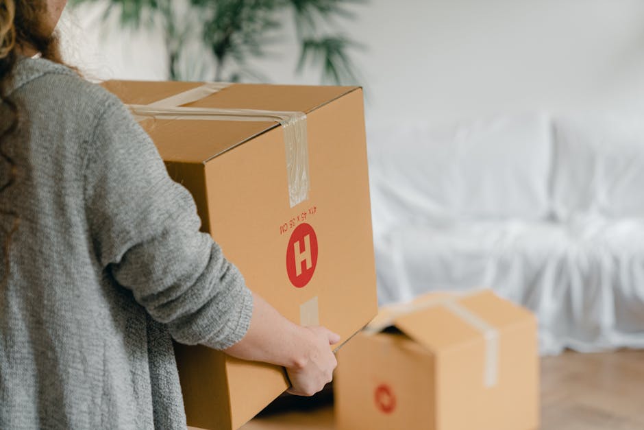A person wearing a grey sweater is seen loading a large cardboard box with a red 'H' logo and product information printed in red onto a moving trolley inside a property. The individual’s hands are visible, gripping the box carefully during the home relocation process. In the background, there is a white upholstered sofa with soft cushions, indicating the living room environment, and a potted plant with green foliage near a window, allowing natural light to brighten the space. The box is sealed with clear packing tape, and additional cardboard boxes are placed on the floor nearby, some also sealed with tape, prepared for transit. The scene depicts a furniture transport and packing stage typical of professional removals, with a focus on safe handling and organized loading, consistent with the services offered by Man with Van The Hale during house removals and moving logistics.