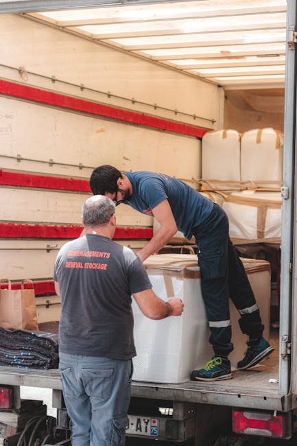 Two movers from Man with Van The Hale are loading large, white foam-padded furniture pieces onto the back of a moving van. One mover, wearing a navy t-shirt and jeans, is standing inside the van, lifting and guiding a sizable wrapped item, while the other, dressed in a grey t-shirt with 'REMOVEMENTS REMOVAL & STORAGE' printed on the back and grey trousers, is outside, assisting with the placement. The interior of the van is visible, with several other boxed or wrapped items stacked against the sides, some secured with brown packing tape. The van's side door is open, revealing the loading area on a paved surface, with a red strip along the interior wall of the van. The environment appears well-lit, and the scene reflects the logistics involved in furniture transport and home relocation facilitated by professional removals services.