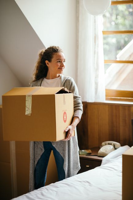 A young couple sitting on a white sofa inside a room with light-colored wooden flooring, surrounded by moving boxes made of brown cardboard with red labels, suggestive of a house relocation process. T