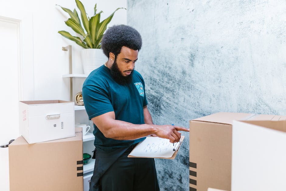 A man with dark curly hair and a beard, dressed in a dark blue t-shirt, is standing inside a room with a textured blue wall behind him. He is holding a clipboard in his left hand and using his right i