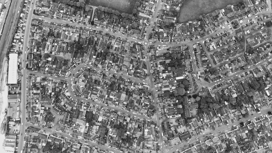 A black-and-white aerial photograph of a dense residential area displaying numerous closely packed houses with varied roof shapes and sizes, arranged along winding and grid-like streets. Narrow alleyways and small gardens are visible between some of the buildings. Larger structures, possibly commercial or community buildings, are located along main roads on the outskirts. Trees and patches of greenery are dispersed throughout the area, providing contrast to the tightly built environment. The image captures the urban layout and infrastructure typical of a suburban neighbourhood, with no visible moving activities, vehicles, or equipment related to house removals or transporting furniture. The setting appears calm and static, reflecting a snapshot of everyday residential life, which might be the context for a moving or house relocation service like Man with Van The Hale, supporting planning for such operations in Hale Barns.