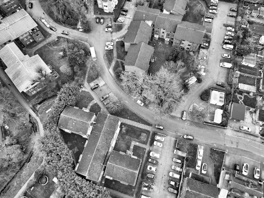 An aerial view of a rural property showing a variety of building structures and outdoor areas. In the centre, a two-storey house with a dark roof is surrounded by a garden with trees, shrubs, and patio furniture. Adjacent to the house, there are several other outbuildings, including a large elongated barn with a light-colored roof and multiple skylights, as well as smaller sheds. A paved driveway runs past the buildings, with several cars parked along it. To the left, a narrow pathway leads to additional structures, while fields of green crops extend to the bottom of the image. The property is part of a rural landscape with open grassy areas and adjacent garden plots, and appears to be in the process of a home relocation involving packing and furniture transport, supported by Man with Van The Hale as shown in the context of house removals and moving logistics.