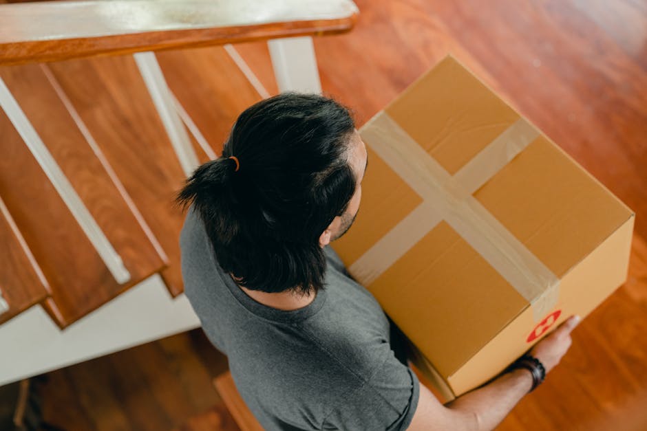 A man with long dark hair tied back, wearing a grey t-shirt, is seen from above carrying a medium-sized cardboard box sealed with clear packing tape and a red recycling symbol on the side. He appears to be inside a home, walking towards a doorway or staircase, as indicated by the wooden floor and a white stair railing visible in the background. The box is carefully held with both hands, suggesting a cautious loading or unloading process typical of house removals and furniture transport. The scene emphasizes the careful handling of packed items during packing and moving operations, supported by the presence of packing materials such as tape, and the indoor environment that is part of a home relocation setting managed by Man with Van The Hale. The lighting is natural and the overall composition highlights the physical effort involved in home relocation tasks.