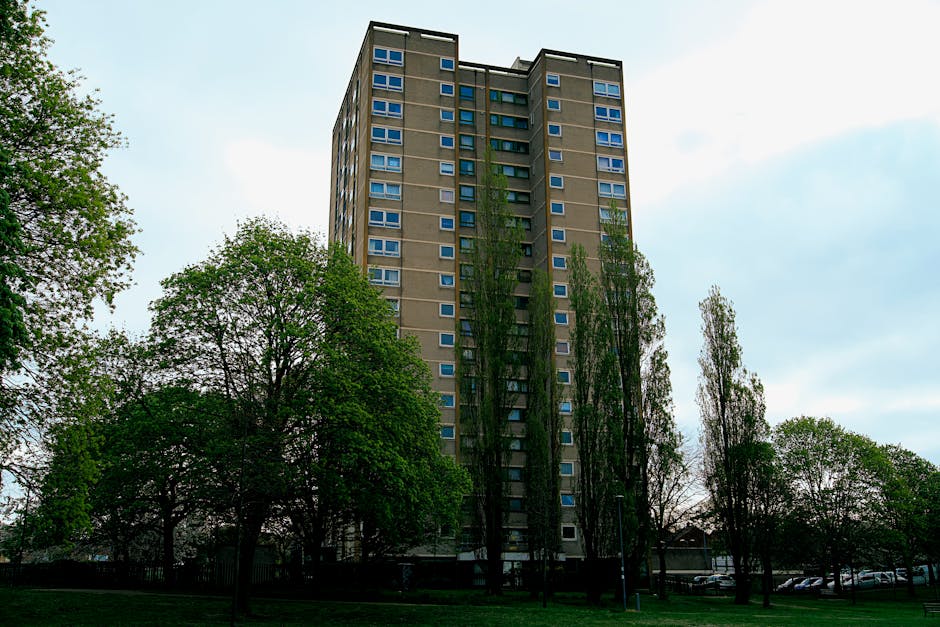 A tall, multi-storey residential building with a modern concrete exterior and numerous uniformly spaced windows, situated in a suburban area with surrounding mature trees and green foliage. The building appears to be part of a housing complex or apartment block, with the trees providing a natural border at the base. The sky above is partly cloudy with patches of blue visible, and the scene is illuminated by natural daylight. This setting relates to house removals and moving services, suggesting a location where furniture transport and home relocation activities may occur, especially in the context of access rules for flats like those in The Hale. Man with Van The Hale offers professional removal and packing services for residents in such high-rise developments, ensuring efficient and secure loadings during the moving process.