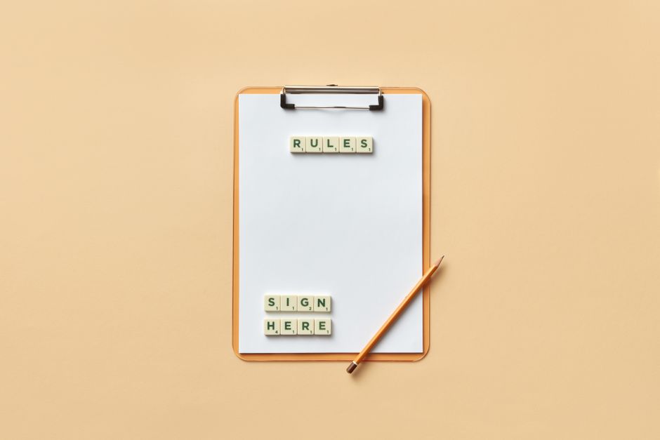 A white clipboard with a wooden frame is positioned vertically on a light beige background. The clipboard holds a blank white sheet of paper, with black clip at the top. On the paper, there are small lettered tiles spelling out 'RULES,' arranged horizontally at the top, and another set of tiles spelling out 'SIGN HERE,' arranged in two rows below. To the right of the clipboard lies a wooden pencil with a sharpened tip, angled slightly upward. The scene is well-lit with soft, even lighting, emphasizing a clean, organized setting that hints at a preparation process for a home relocation or moving instructions. The presence of the clipboard and sign tiles subtly relates to move-in or move-out procedures, fitting with services offered by Man with Van The Hale, connected to house removals and furniture transport in The Hale flats.