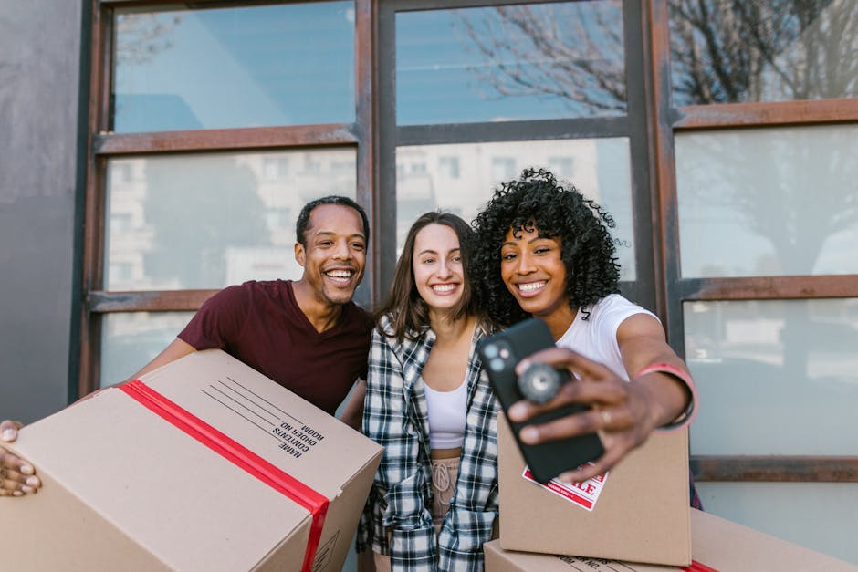 Three smiling individuals standing outside a house with large glass windows, holding cardboard boxes sealed with red tape. The person on the right, an African-American woman with curly hair, is taking a selfie with a smartphone while holding a box. The man on the left, of African descent, and the woman in the middle, with light skin and straight brown hair, are also holding boxes. The boxes are labeled with packing and moving materials, suggesting a home relocation or furniture transport process. Behind them, the house's exterior features wooden framing and a reflection of nearby buildings can be seen in the windows. This scene illustrates aspects of packing and moving logistics as part of house removals, aligning with services offered by [COMPANY_NAME] for local house relocations in Hale Village, emphasizing seamless packing, loading, and transport activities.