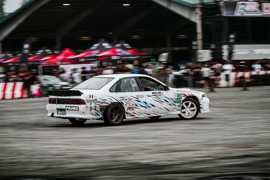 A white racing car with a colourful blue, red, and black stripe design is captured in motion on a tarmac surface, likely during a motorsport event in an urban environment. The vehicle has various stickers and decals, including the word 'PANIC' on the rear bumper and other promotional graphics on its sides. Behind the car, there are blurred figures of spectators, as well as structures with red and black tents and a bridge or elevated roadway overhead, indicating a busy outdoor setting. The scene suggests a dynamic environment, possibly related to vehicle transport or an event involving moving vehicles within a city. Although the focus is on rapid movement, the context hints at logistical aspects of vehicle manoeuvres, which parallels the themes of house removals and furniture transport handled by [COMPANY_NAME] in similar settings.