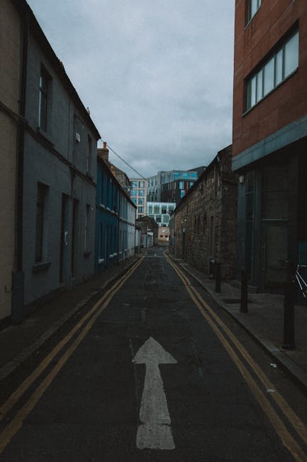 A narrow city street in Bowdon, featuring adjacent buildings on both sides with contrasting architectural styles; on the left, older, weathered buildings with dark-colored facades and small windows, while on the right, a modern, multi-storey brick building with large windows. The street is paved with asphalt, marked by a single white arrow painted on the surface indicating traffic direction, and flanked by black bollards on the right sidewalk. Overhead, a cloudy grey sky creates an overcast atmosphere. In the distance, there are more modern office or apartment buildings with glass facades. The street appears quiet, possibly during a relocation or moving project, with no vehicles or pedestrians visible. This scene supports the context of furniture manoeuvres and home relocation navigating narrow streets, relevant to the services offered by Man with Van The Hale in house removals and transport solutions.
