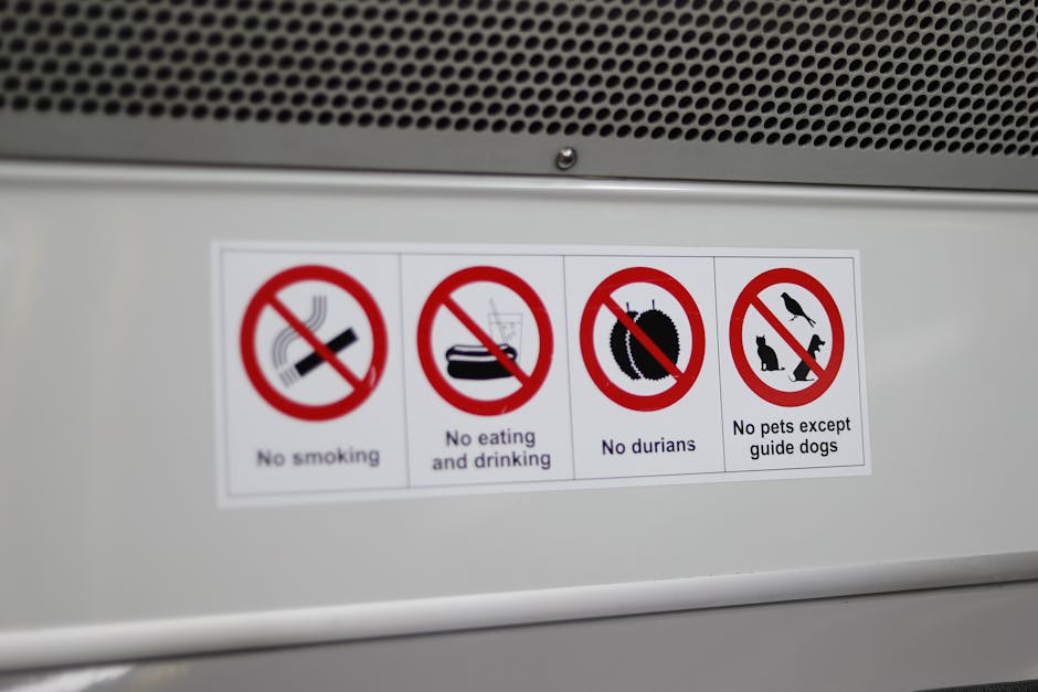 Close-up of a white label affixed to a metallic surface, displaying four icons with red prohibition symbols and accompanying text, indicating restrictions such as no smoking, no eating or drinking, no durians, and no pets except guide dogs. The label is positioned near a vent or grille at the top of the image, inside a property or vehicle used for home relocation services by Man with Van The Hale. The surrounding environment includes a smooth metallic surface and part of the ventilation area, with the lighting suggesting an indoor or enclosed space. This image illustrates safety and regulatory instructions relevant to furniture transport and packing processes involved in an internal or external house removal, supporting logistical considerations for moving companies.