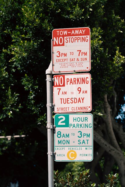 Close-up view of a multi-panel parking and parking restriction signs mounted on a metal pole outdoors, with a background of lush green trees and bright blue sky. The top panel is red and indicates a tow-away zone with no stopping permitted from 3 pm to 7 pm, except on Saturdays and Sundays, with additional lockdown and contact details. The middle panel, also red, states no parking from 7 am to 9 am on Tuesdays, noting street cleaning restrictions. The bottom panel has a green section at the top with white text indicating two-hour parking restrictions from 8 am to 3 pm, Monday to Saturday, with an exception for vehicles with area C permits, indicated by a yellow circle symbol. The signs are positioned to inform residents and visitors of local parking rules near Hale Station, relevant to home relocation or moving logistics, as handled by Man with Van The Hale.