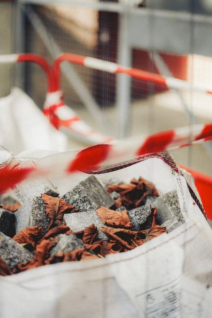 Close-up of a white woven polypropylene sack filled with crushed stone and scattered dry brown leaves, situated on a paved surface near a building under construction or renovation. Behind the sack, th