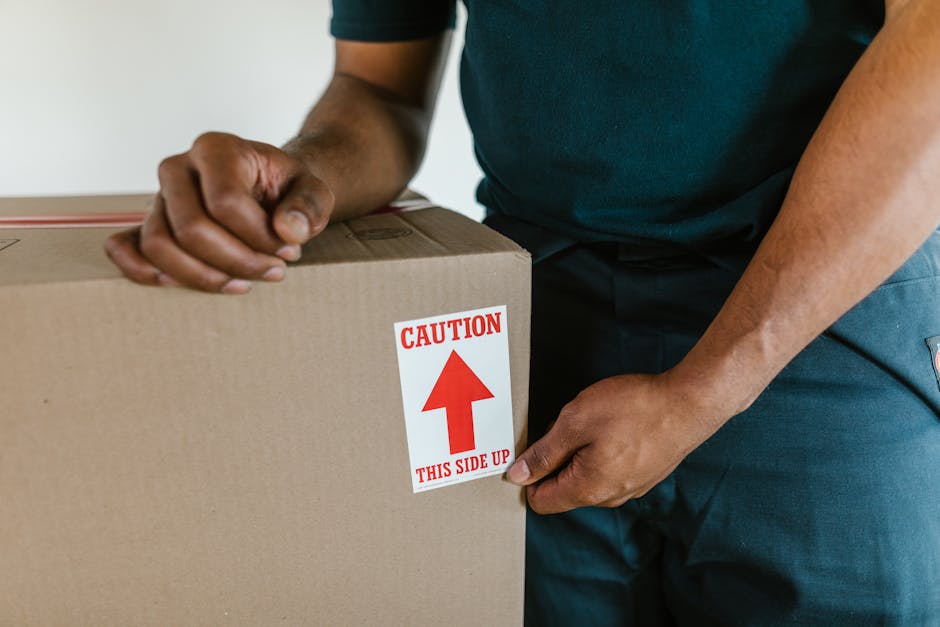 A close-up of a person wearing a dark teal shirt and dark trousers, securing a cardboard moving box with a label that reads 'CAUTION THIS SIDE UP' with a red arrow pointing upwards. The individual is indoors, near a doorway, preparing the box for home relocation or furniture transport as part of packing and moving procedures. The box is made of brown corrugated cardboard, and the person is holding the box steady while ensuring the label is visible, indicating careful handling during the loading process. In the background, a partially visible van or vehicle may be nearby, suggesting readiness for transport. This scene illustrates the packing and loading phase of a house removal service offered by Man with Van The Hale, highlighting the importance of proper packaging, safety labels, and careful handling during a residential moving operation.