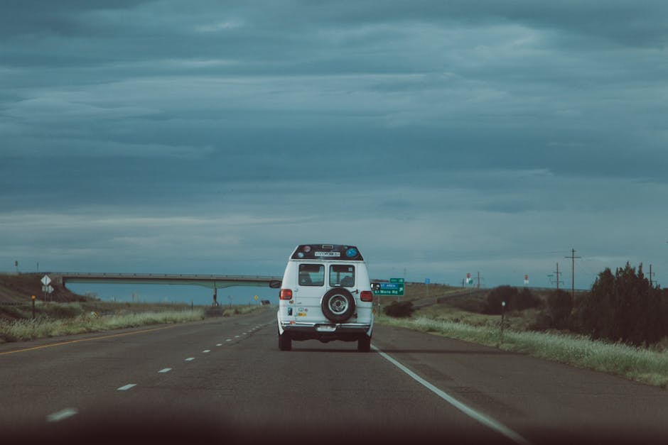 A white van with a spare tyre mounted on the rear driving along an open, two-lane road with dashed white lane markings, under a dark, cloudy sky. The vehicle is positioned in the middle of the lane, heading away from the camera. The landscape on either side of the road features grassy shoulders and distant trees, with a bridge visible overhead and a road sign in the background indicating directions. This scene captures the typical environment associated with home relocation or furniture transport as part of a professional removals service, such as those provided by Man with Van The Hale. The image emphasizes the transportation process involved in packing and moving, illustrating a key stage of house removals in progress.