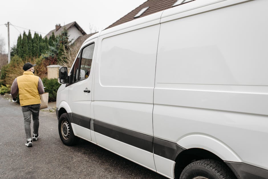 A person wearing a yellow puffer vest, black cap, and grey joggers is walking towards the side of a large white delivery van parked on Ashley Road, Hale, with residential houses and greenery in the background. The van is positioned near a curb on a paved street, with its side door closed. The scene captures the loading or unloading process associated with house removals, as the individual appears to be handling packing materials or belongings in preparation for a home relocation. The white van belongs to Man with Van The Hale, a company specializing in removals, and is part of the furniture transport and packing and moving tasks typical of professional house removal services. The environment is lit by natural daylight, and the overall setting suggests a residential area suitable for local or regional moving logistics.